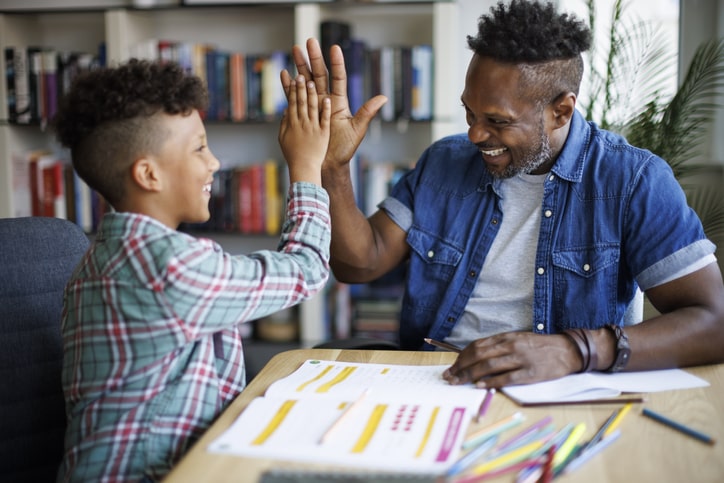 Dad helps son with homework. They are both happy and high-five. Report card advice for parents: make learning fun. 